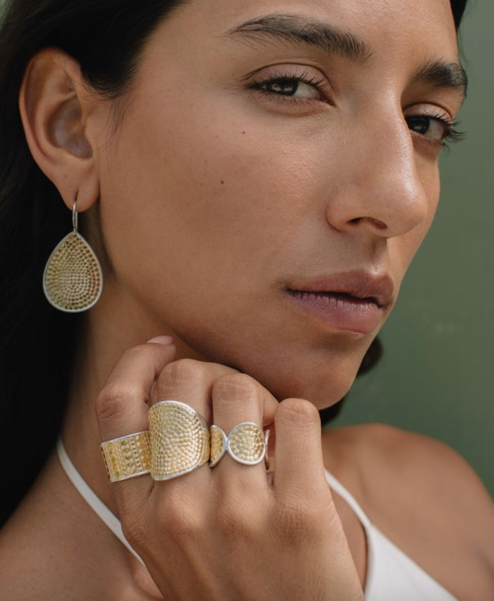 Woman wearing gold earrings and ring against a neutral background from Anna Beck