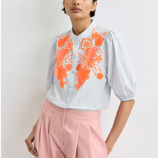 Woman wearing a pale blue blouse with orange floral embroidery and pink pants on a light background