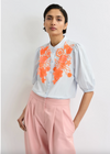 Woman wearing a pale blue blouse with orange floral embroidery and pink pants on a light background