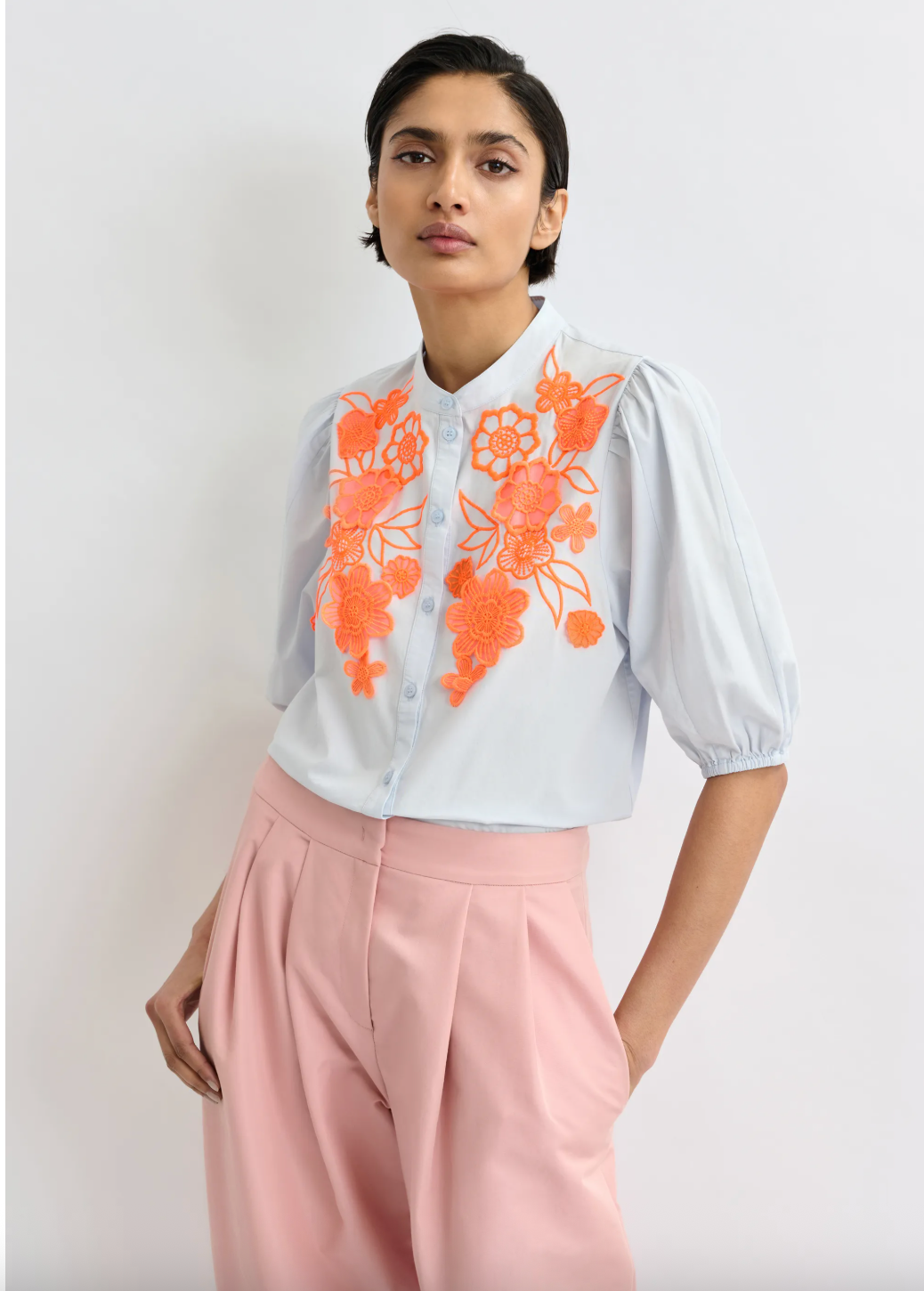 Woman wearing a pale blue blouse with orange floral embroidery and pink pants on a light background