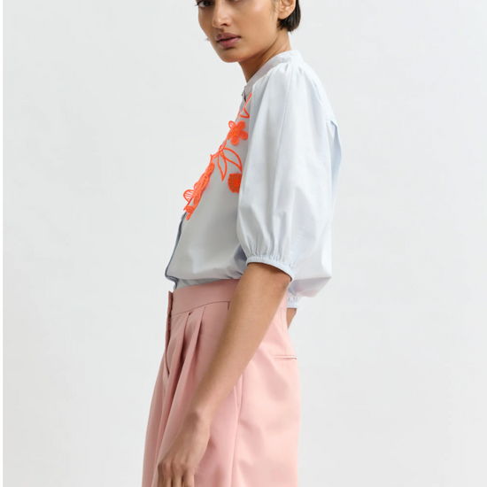 Woman wearing a pale blue blouse with orange design and pink pants on a light gray background