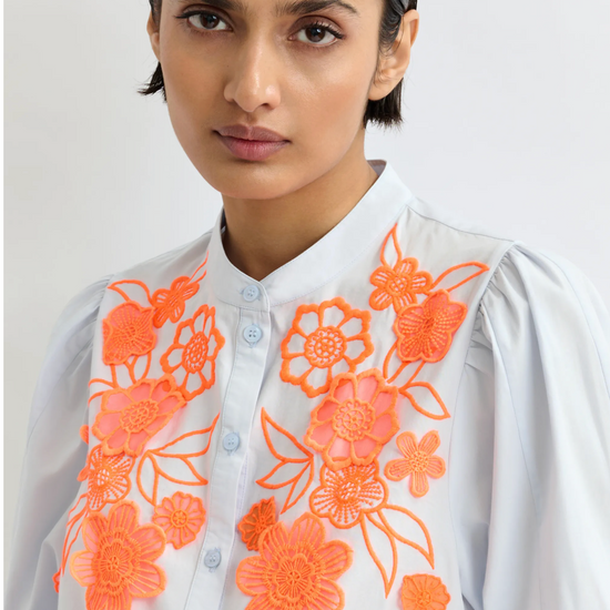 Woman wearing a pale blue shirt with orange floral embroidery on a plain background