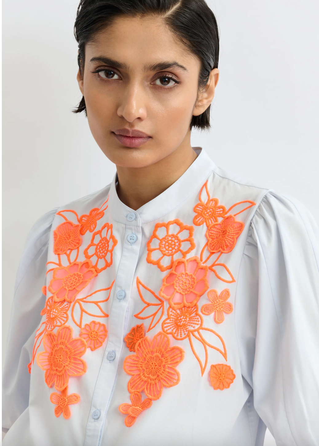 Woman wearing a pale blue shirt with orange floral embroidery on a plain background