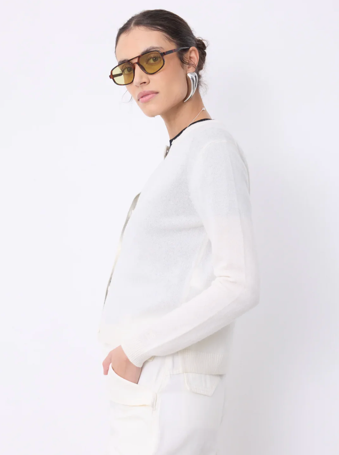 A model wearing a white knit long-sleeve cardigan and matching white trousers, styled with yellow-tinted sunglasses and silver hoop earrings against a plain white background.
