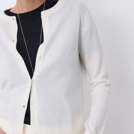 A close-up of a person wearing a cream-colored knit cardigan over a black top, with a long silver necklace visible against a plain light background.