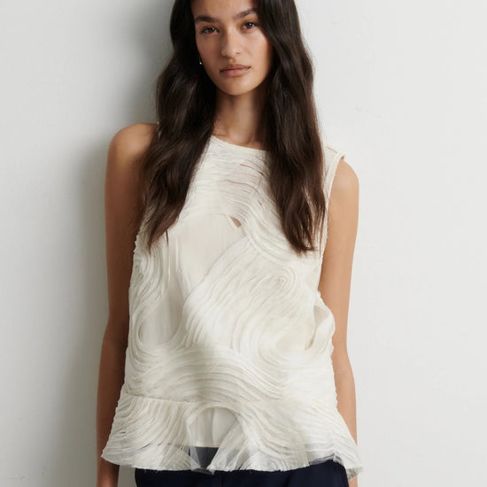 A woman wearing a sleeveless textured white top with a wavy, pleated design and a ruffled hem, photographed against a plain light gray wall.