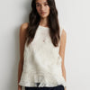 A woman wearing a sleeveless textured white top with a wavy, pleated design and a ruffled hem, photographed against a plain light gray wall.