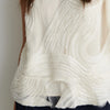 Close-up of a sleeveless white top with dramatic layered ruffles and textured pleating, worn by a model.