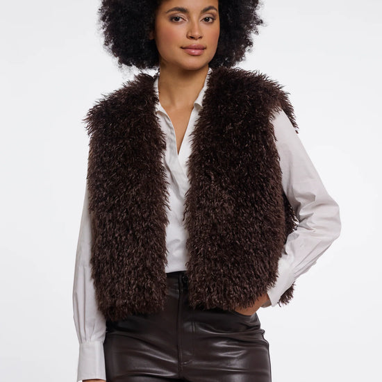 A model with an afro hairstyle wearing a dark brown curly faux fur vest over a white button-down shirt and dark brown faux leather pants, photographed against a solid white studio background.