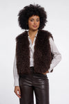 A model with an afro hairstyle wearing a dark brown curly faux fur vest over a white button-down shirt and dark brown faux leather pants, photographed against a solid white studio background.