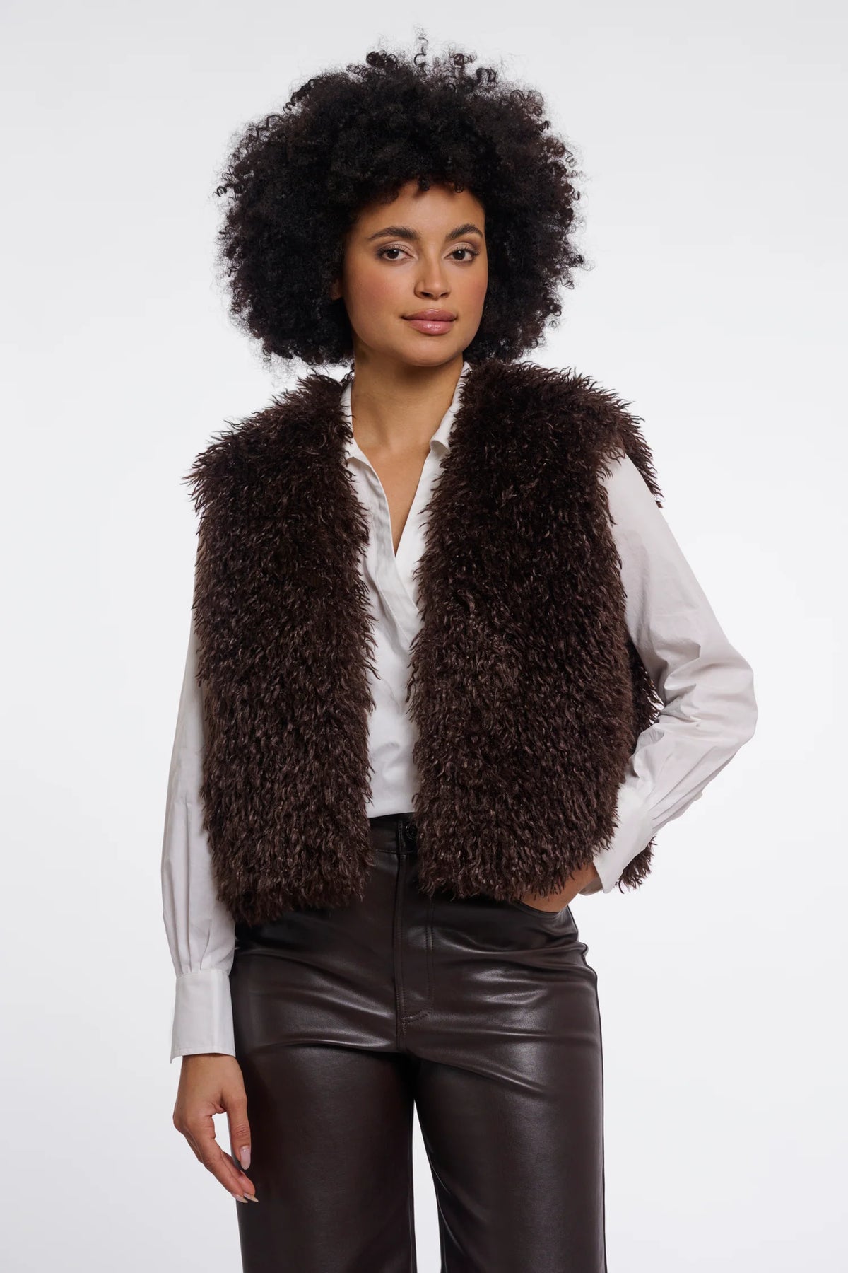 A model with an afro hairstyle wearing a dark brown curly faux fur vest over a white button-down shirt and dark brown faux leather pants, photographed against a solid white studio background.