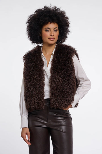 A model with an afro hairstyle wearing a dark brown curly faux fur vest over a white button-down shirt and dark brown faux leather pants, photographed against a solid white studio background.