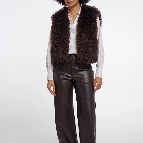 A model with an afro hairstyle stands against a light gray studio background wearing a dark brown shaggy faux-fur vest over a white button-up shirt, paired with dark brown faux-leather wide-leg pants and black pointed shoes.