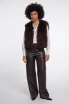 A model with an afro hairstyle stands against a light gray studio background wearing a dark brown shaggy faux-fur vest over a white button-up shirt, paired with dark brown faux-leather wide-leg pants and black pointed shoes.