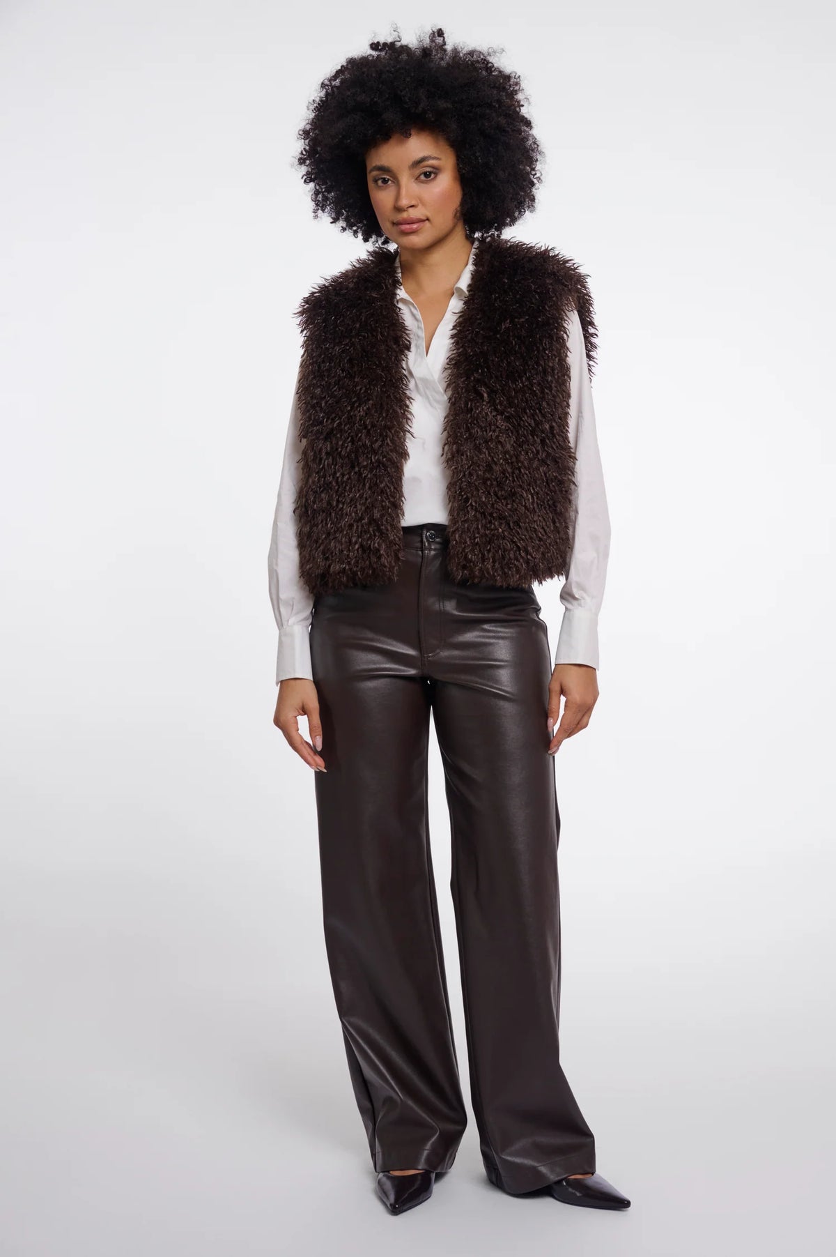 A model with an afro hairstyle stands against a light gray studio background wearing a dark brown shaggy faux-fur vest over a white button-up shirt, paired with dark brown faux-leather wide-leg pants and black pointed shoes.