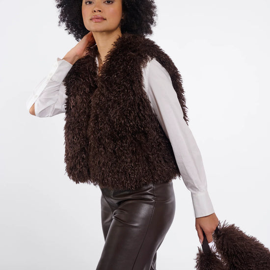A model with an afro hairstyle is wearing a dark brown curly faux fur vest over a white long-sleeve shirt and dark brown faux leather pants, holding a matching brown faux fur bag against a light gray studio background.