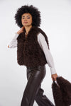 A model with an afro hairstyle is wearing a dark brown curly faux fur vest over a white long-sleeve shirt and dark brown faux leather pants, holding a matching brown faux fur bag against a light gray studio background.