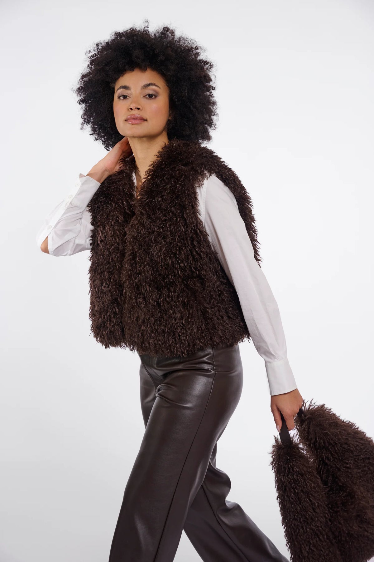 A model with an afro hairstyle is wearing a dark brown curly faux fur vest over a white long-sleeve shirt and dark brown faux leather pants, holding a matching brown faux fur bag against a light gray studio background.