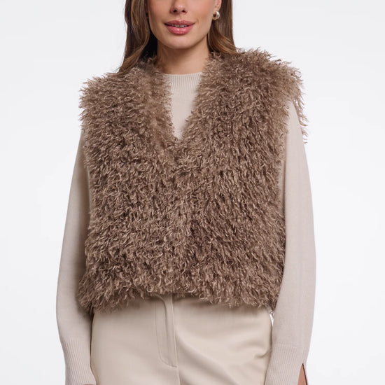 A woman modeling a shaggy, textured brown faux fur vest over a cream long-sleeve top and cream trousers, set against a plain light gray studio background.