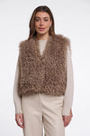 A woman modeling a shaggy, textured brown faux fur vest over a cream long-sleeve top and cream trousers, set against a plain light gray studio background.