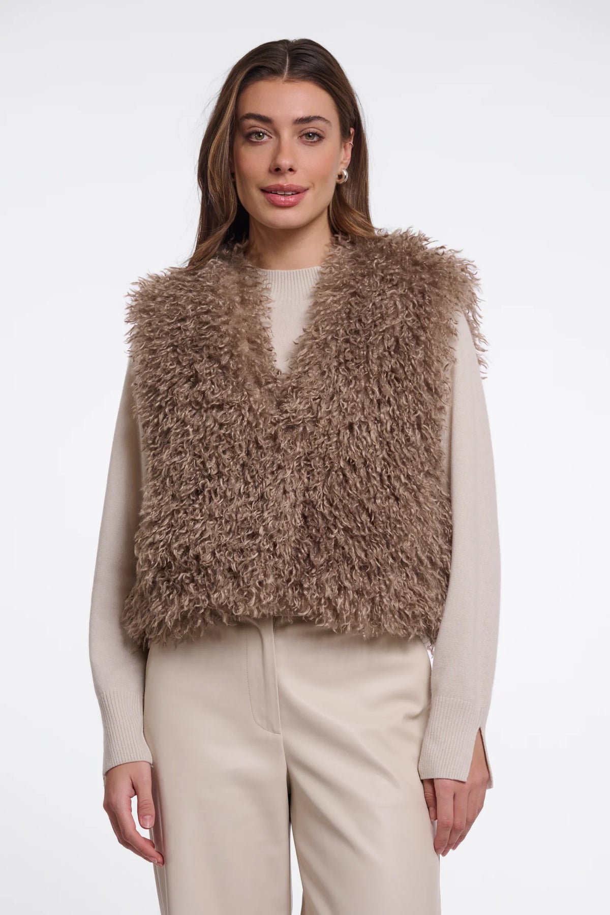 A woman modeling a shaggy, textured brown faux fur vest over a cream long-sleeve top and cream trousers, set against a plain light gray studio background.