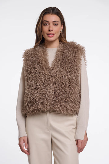 A woman modeling a shaggy, textured brown faux fur vest over a cream long-sleeve top and cream trousers, set against a plain light gray studio background.