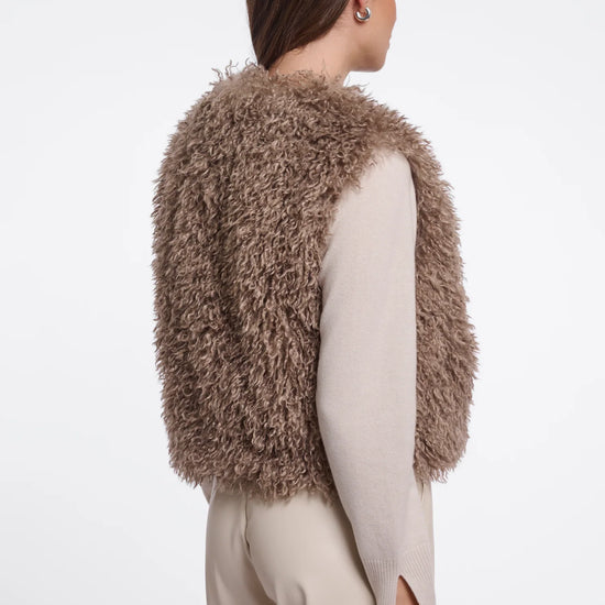 A woman shown from the back wearing a shaggy, cropped taupe-brown vest over a light beige long-sleeve top and beige trousers, against a plain white studio background.