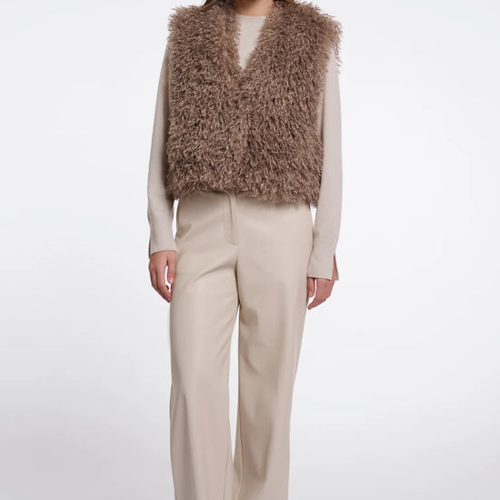 A model stands against a light gray studio background wearing a shaggy taupe-brown faux fur vest over a cream long-sleeve top and beige wide-leg trousers.