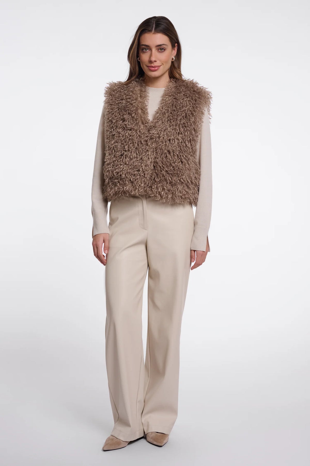 A model stands against a light gray studio background wearing a shaggy taupe-brown faux fur vest over a cream long-sleeve top and beige wide-leg trousers.