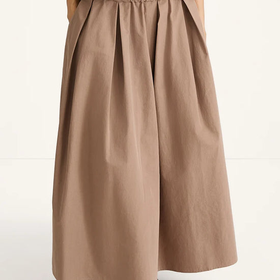 A person wearing a wide, pleated maxi skirt in a warm taupe-brown color, paired with a fuzzy brown top and dark brown flat shoes, photographed against a clean white studio background.