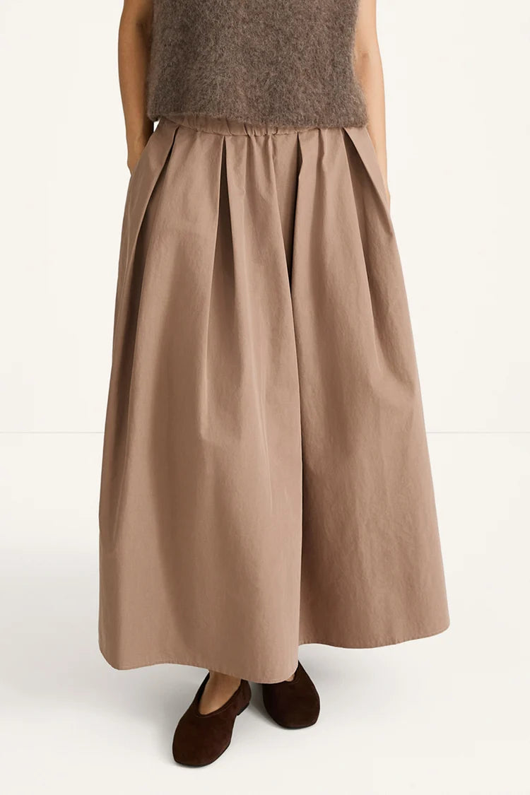 A person wearing a wide, pleated maxi skirt in a warm taupe-brown color, paired with a fuzzy brown top and dark brown flat shoes, photographed against a clean white studio background.