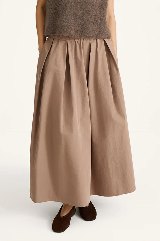 A person wearing a wide, pleated maxi skirt in a warm taupe-brown color, paired with a fuzzy brown top and dark brown flat shoes, photographed against a clean white studio background.