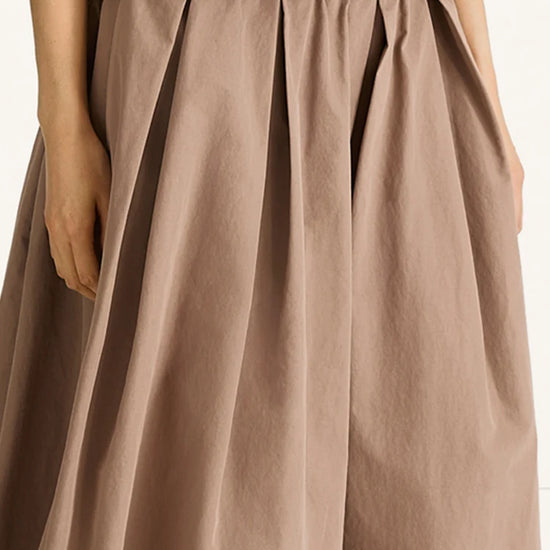 A close-up of a pleated, wide-leg skirt in a muted taupe-brown color, worn by a model with a fuzzy brown top partially visible at the top edge.