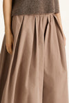 A close-up of a pleated, wide-leg skirt in a muted taupe-brown color, worn by a model with a fuzzy brown top partially visible at the top edge.