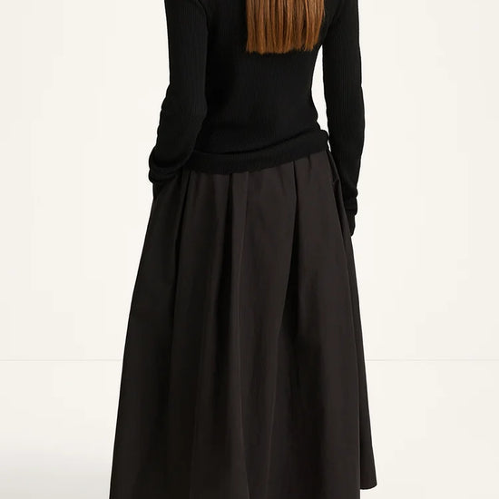 A woman shown from the back wearing a black long-sleeve mock-neck top and a black pleated midi skirt, paired with black shoes, against a plain white studio background.