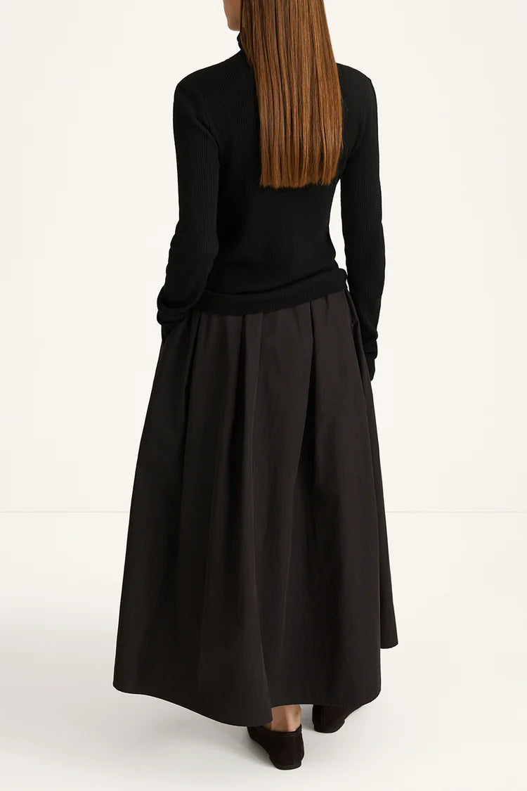 A woman shown from the back wearing a black long-sleeve mock-neck top and a black pleated midi skirt, paired with black shoes, against a plain white studio background.