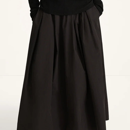 A person wearing a long black pleated skirt with a black ribbed long-sleeve top and black slip-on shoes, photographed against a plain light background.