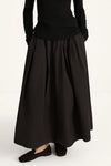 A person wearing a long black pleated skirt with a black ribbed long-sleeve top and black slip-on shoes, photographed against a plain light background.