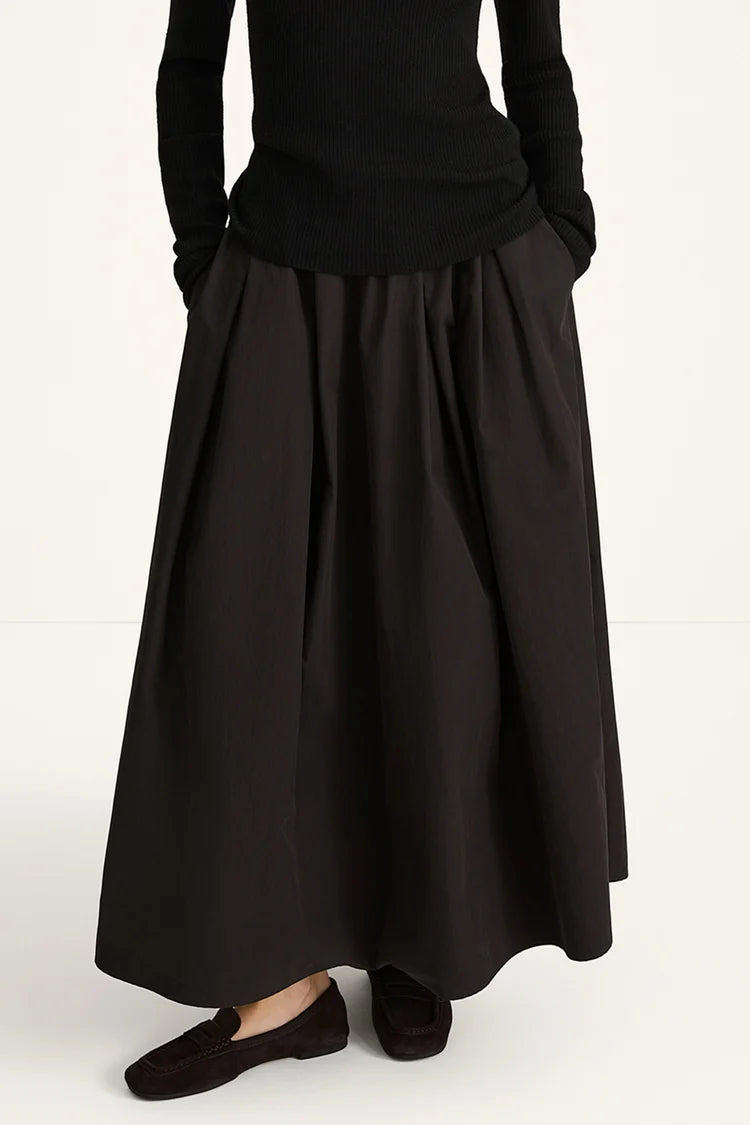 A person wearing a long black pleated skirt with a black ribbed long-sleeve top and black slip-on shoes, photographed against a plain light background.