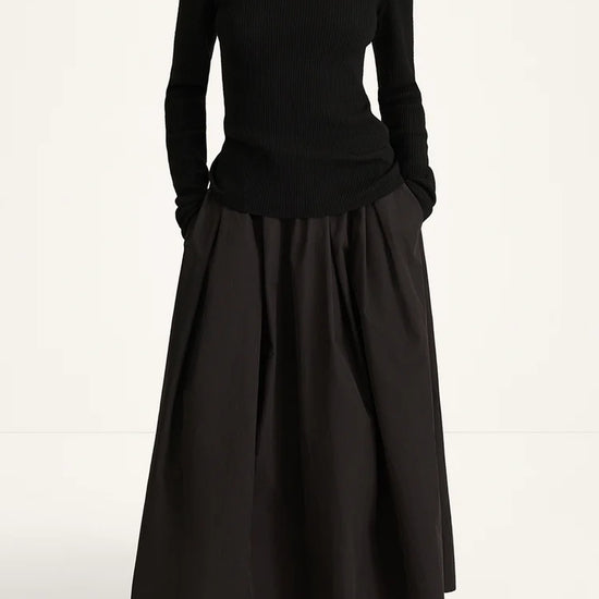 A model wearing a black long-sleeve turtleneck sweater and a matching black pleated maxi skirt, paired with black flat shoes, against a plain white studio background.