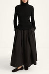 A model wearing a black long-sleeve turtleneck sweater and a matching black pleated maxi skirt, paired with black flat shoes, against a plain white studio background.