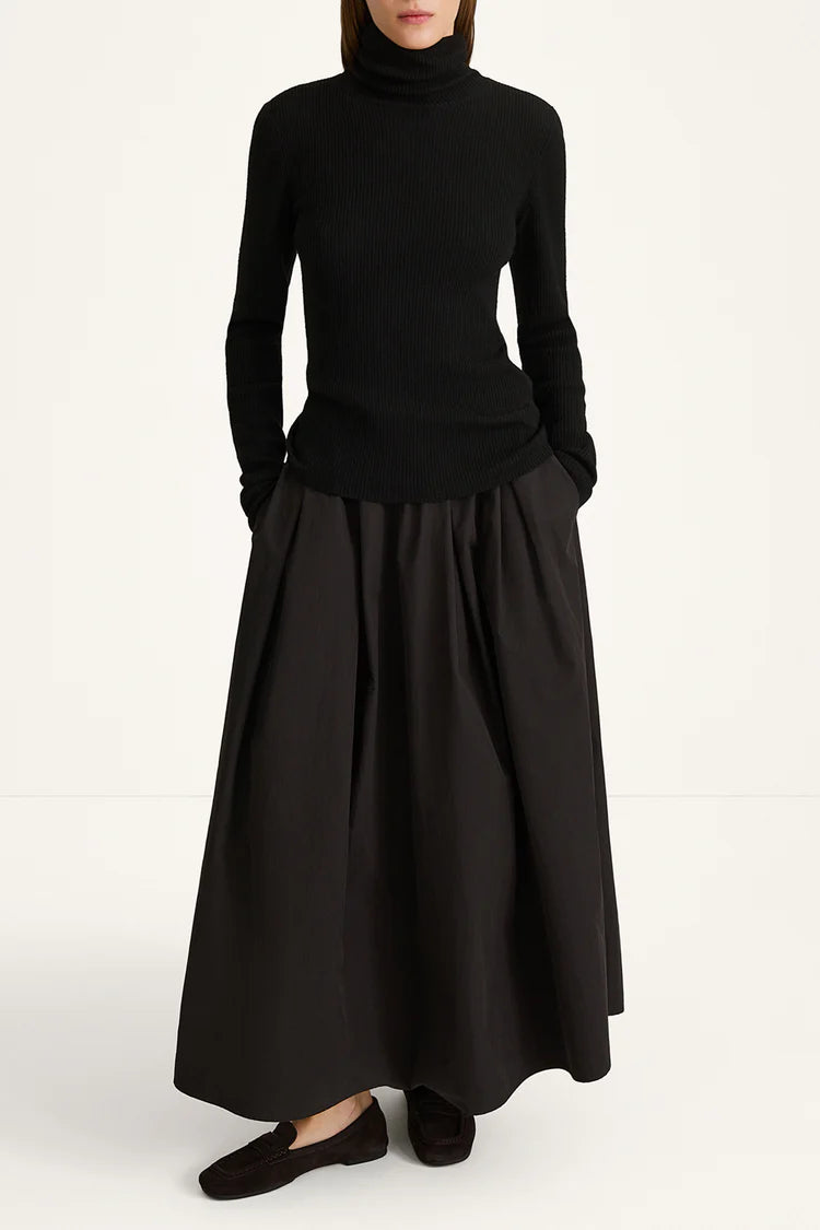 A model wearing a black long-sleeve turtleneck sweater and a matching black pleated maxi skirt, paired with black flat shoes, against a plain white studio background.