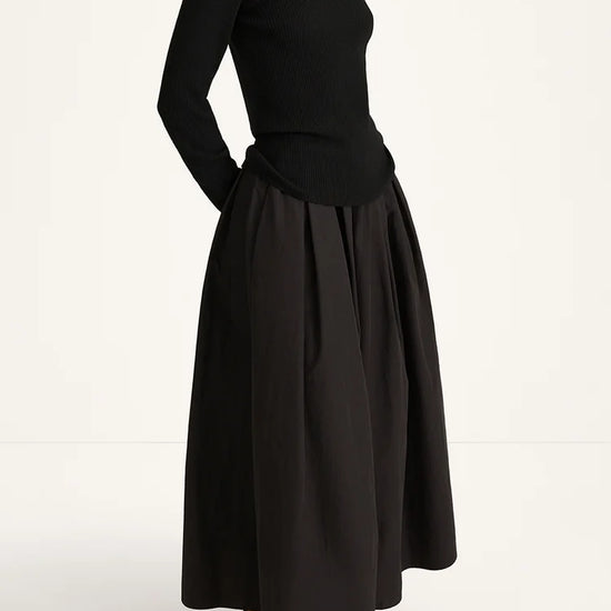 A model wearing a black long-sleeve turtleneck top and a matching black pleated maxi skirt, paired with black loafers, photographed against a clean white studio background.