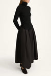 A model wearing a black long-sleeve turtleneck top and a matching black pleated maxi skirt, paired with black loafers, photographed against a clean white studio background.