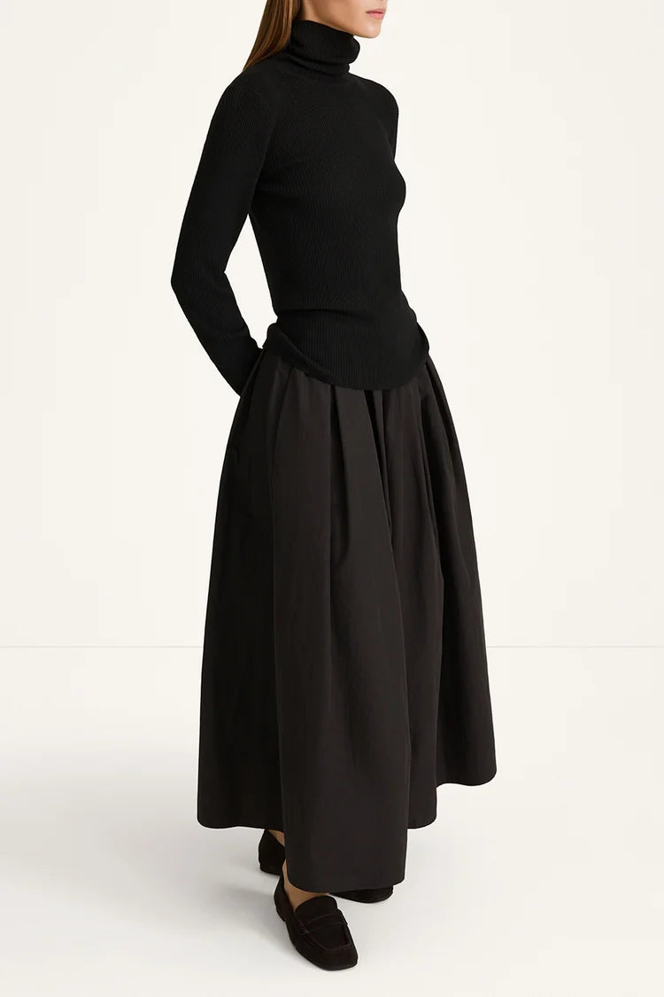 A model wearing a black long-sleeve turtleneck top and a matching black pleated maxi skirt, paired with black loafers, photographed against a clean white studio background.