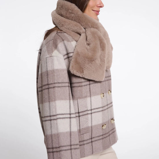 A woman shown in side profile wearing a beige and brown plaid double-breasted coat with a thick, plush tan faux-fur collar, paired with light beige trousers, against a plain white studio background.