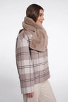A woman shown in side profile wearing a beige and brown plaid double-breasted coat with a thick, plush tan faux-fur collar, paired with light beige trousers, against a plain white studio background.