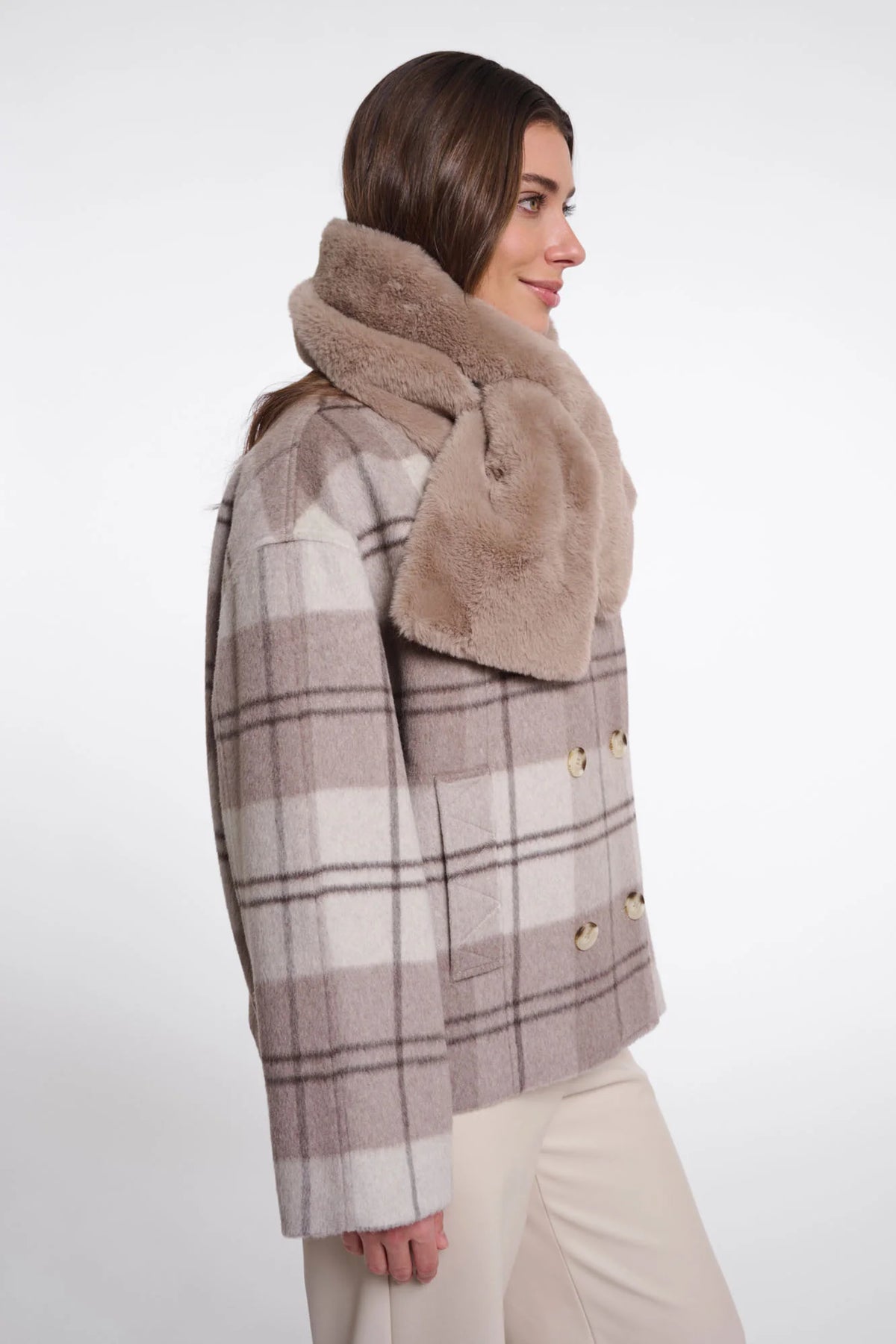 A woman shown in side profile wearing a beige and brown plaid double-breasted coat with a thick, plush tan faux-fur collar, paired with light beige trousers, against a plain white studio background.