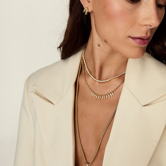 A close-up of a woman wearing layered gold necklaces, including a delicate chain necklace with a round pendant, a spiky spike necklace, and a pearl-like chain, paired with a cream blazer.