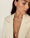 A close-up of a woman wearing layered gold necklaces, including a delicate chain necklace with a round pendant, a spiky spike necklace, and a pearl-like chain, paired with a cream blazer.